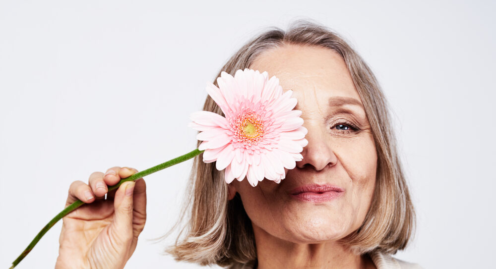 Smiling older woman holding a pink gerbera flower over one eye