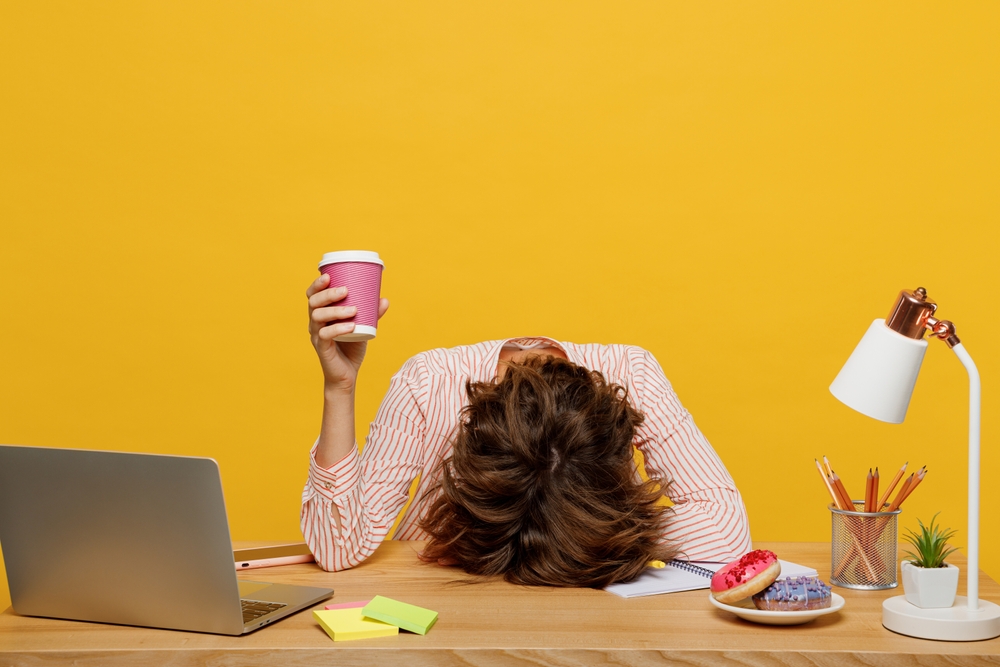 Exhausted worker slumped on desk holding coffee, illustrating workplace stress and burnout